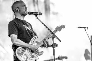 Male guitarist singing into a microphone while playing a sticker-covered electric guitar on stage in black-and-white.