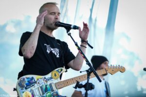 Male guitarist singing into a microphone with eyes closed during an outdoor performance; guitar covered in stickers.