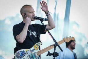Male guitarist and vocalist performing on stage, singing into a microphone with his left hand raised in a live concert setting.