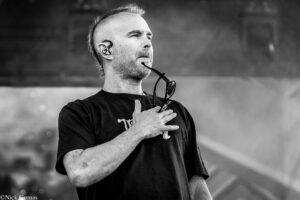 Black-and-white photo of a man performing on stage, with in-ear monitors, holding sunglasses near his mouth while looking upward.