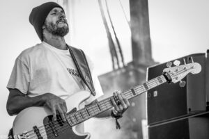 Bearded bass guitarist wearing a knit beanie playing a bass on stage.