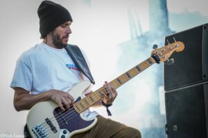 Male guitarist wearing a black beanie and white T-shirt, playing a white Fender bass on stage with a large speaker cabinet nearby.