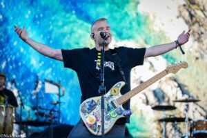 Male rock guitarist singing into a microphone with arms spread wide on stage, colorful blue-green backdrop behind him, guitar covered in stickers.