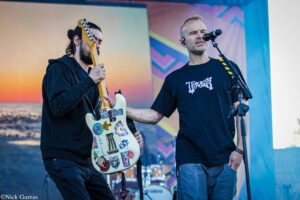Two musicians on stage: a guitarist with a sticker-covered white guitar, and a singer into a microphone.