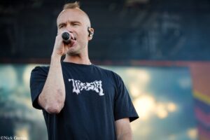 Male performer singing into a handheld microphone on a dimly lit stage, wearing a black T-shirt with graphics in a crowd-friendly setting.