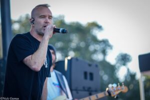 Male singer performing on an outdoor stage, holding a microphone with a guitar in the foreground and blurred crowd backdrop menacingly out of focus (in-ear monitor visible).