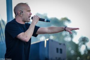 Male singer on an outdoor stage, holding a microphone and extending an arm toward the audience.