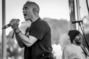 Male singer on stage, gripping a microphone with an in-ear monitor in his ear, performing outdoors (black and white).