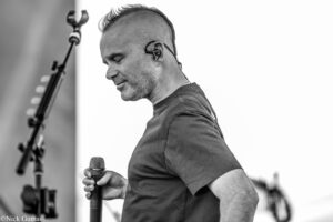 Male performer in a T-shirt on stage, holding a microphone, wearing in-ear monitors and a neck chain, captured in profile in black and white.