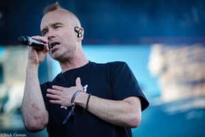 Male singer performs into a handheld microphone on stage, wearing a black T-shirt and wristbands.