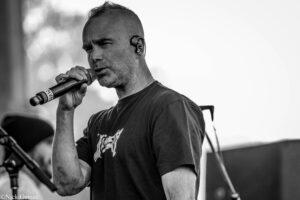 Male singer wearing in-ear monitors, holding a microphone close to his mouth on stage in black and white.