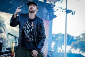 Male singer in a black leather jacket points toward the audience while performing on an outdoor stage.