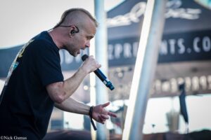 Male singer performing live, mic close to his mouth, wearing in-ear monitor, on stage with blurred backdrop behind him.
