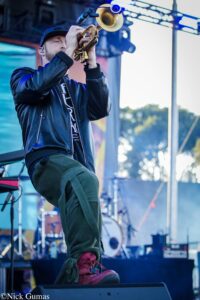 Trumpet player on stage wearing a black leather jacket and cap, intensely blowing into the brass horn with stage lights in the background.