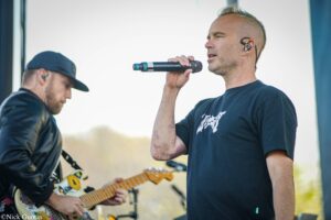 Two musicians perform on an outdoor stage: a singer with in-ear monitors sings into a handheld mic while a guitarist in a cap plays a decorated electric guitar in the background.