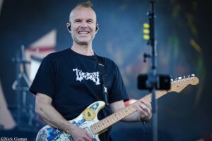 Smiling guitarist playing a cream-colored electric guitar on stage, wearing a black T-shirt and in-ear monitors.