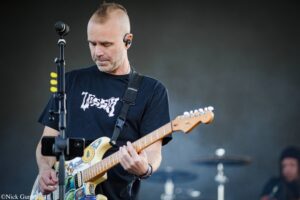 Guitarist in a black T-shirt playing a sticker-covered electric guitar on stage, microphone visible nearby.