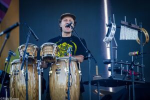 Male drummer wearing a cap sings into a microphone while playing four wooden conga drums on stage with percussion gear nearby.