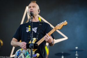 Male guitarist singing into a microphone on stage, wearing a black T-shirt and playing a decorated electric guitar.