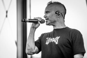 Male vocalist mid-performance, singing into a handheld microphone on stage, with an in-ear monitor visible in his ear.