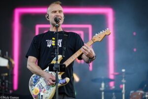 Male guitarist singing into a microphone on stage, wearing a black T-shirt and playing a decorated electric guitar with numerous stickers; pink neon backdrop in the background.