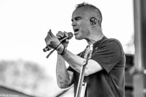Male guitarist and vocalist performing on stage, singing into a microphone with in-ear monitor in place and a dark T-shirt (black-and-white photo).