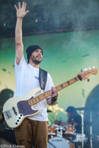 Bassist with cream-colored guitar raises right hand on stage, wearing a black beanie and white t-shirt against a green-yellow backdrop.