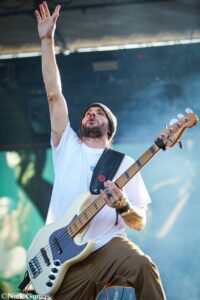 Bearded bassist on stage raising his left arm while playing a cream-colored bass guitar outdoors