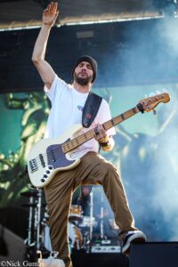 Bass guitarist wearing a white shirt and beanie raises his hand on an outdoor stage.