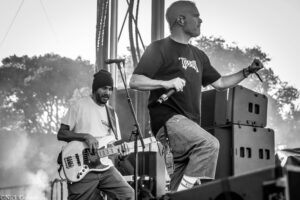 Black-and-white photo of two musicians on an outdoor stage: a lead singer with a microphone, and a bassist playing bass guitar behind him under stage equipment.