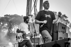 Two musicians perform on an outdoor stage: a singer in a dark shirt front and center, and a guitarist behind him. (black and white)
