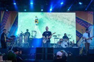Band on stage performing live; guitarist center, drummer behind, keyboardist left; large LED screen behind shows a green image.