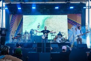 Band performs on an outdoor stage with a large LED screen behind them; lead singer with guitar at center, drummer and other musicians visible, audience in foreground wearing hats.