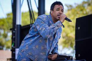 Male singer wearing a floral denim shirt, performing into a handheld microphone on an outdoor stage with a blurred green background.