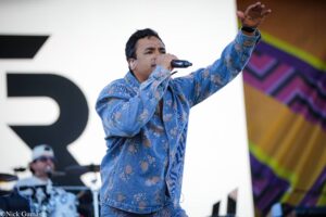 Male singer performing on stage, microphone in hand, wearing a blue floral shirt and raising his arm to engage the crowd