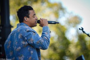 Male singer in a blue floral denim jacket performs outdoors, microphone near his mouth with in-ear monitor visible in ear.