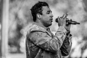 Male singer performing on stage, holding a microphone close to his mouth, wearing a patterned denim jacket (black and white).