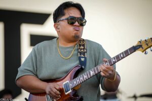 Guitarist playing a sunburst electric guitar outdoors, wearing sunglasses and a gold chain.