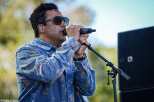 Male singer wearing sunglasses and a floral denim shirt, singing into a microphone outdoors with a monitor screen nearby.