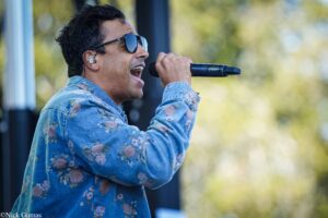 Male singer wearing sunglasses and a floral denim jacket, singing into a wireless handheld microphone outdoors.