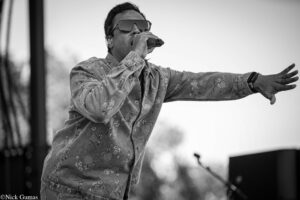 Male singer wearing sunglasses and a floral denim jacket, performing into a microphone with outstretched arm on stage (black and white).