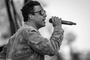 Male singer wearing sunglasses and a floral denim jacket, singing into a handheld microphone.
