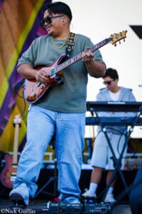 Two musicians on stage: a man in a green shirt and sunglasses playing a brown electric guitar, with a keyboardist in the background.