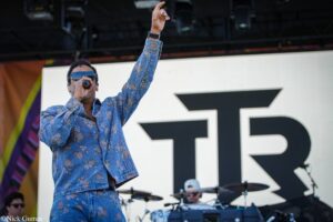 Male singer on stage raising his right hand, wearing a blue floral denim shirt, performing in front of a large screen backdrop.