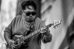 Guitarist wearing sunglasses plays an electric guitar on stage, close-up of upper body and instrument in black and white