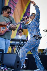 Two musicians perform on stage: a guitarist in green shirt and sunglasses playing a wooden-orange guitar, while a singer in a floral denim suit belts into a microphone wearing sunglasses.