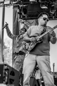 Two male musicians performing on stage: a guitarist wearing sunglasses and a chain plays an electric guitar while a singer behind him raises an arm and belts out a note.