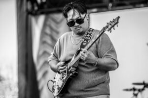 Black-and-white photo of a man wearing sunglasses, playing an electric guitar on stage.