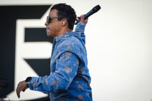 Male performer in a floral denim shirt wearing sunglasses, holding a microphone behind his head while on stage.