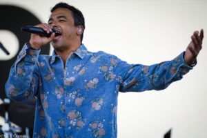 Male singer in a blue floral shirt sings into a microphone with one arm raised on stage.